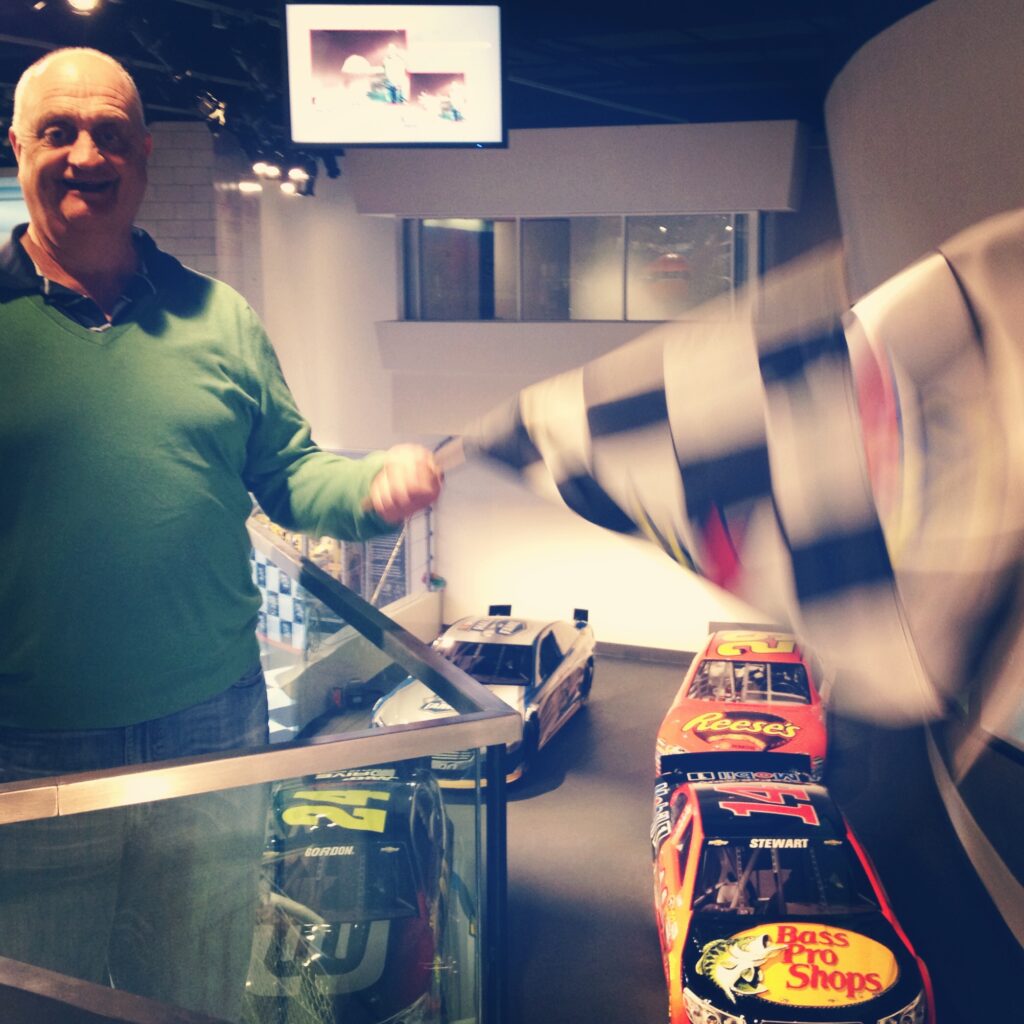 A smiling man in a green sweater waves a checkered flag on an indoor balcony above three NASCAR race cars—one of the top things to do in Charlotte—with a TV screen visible in the background.