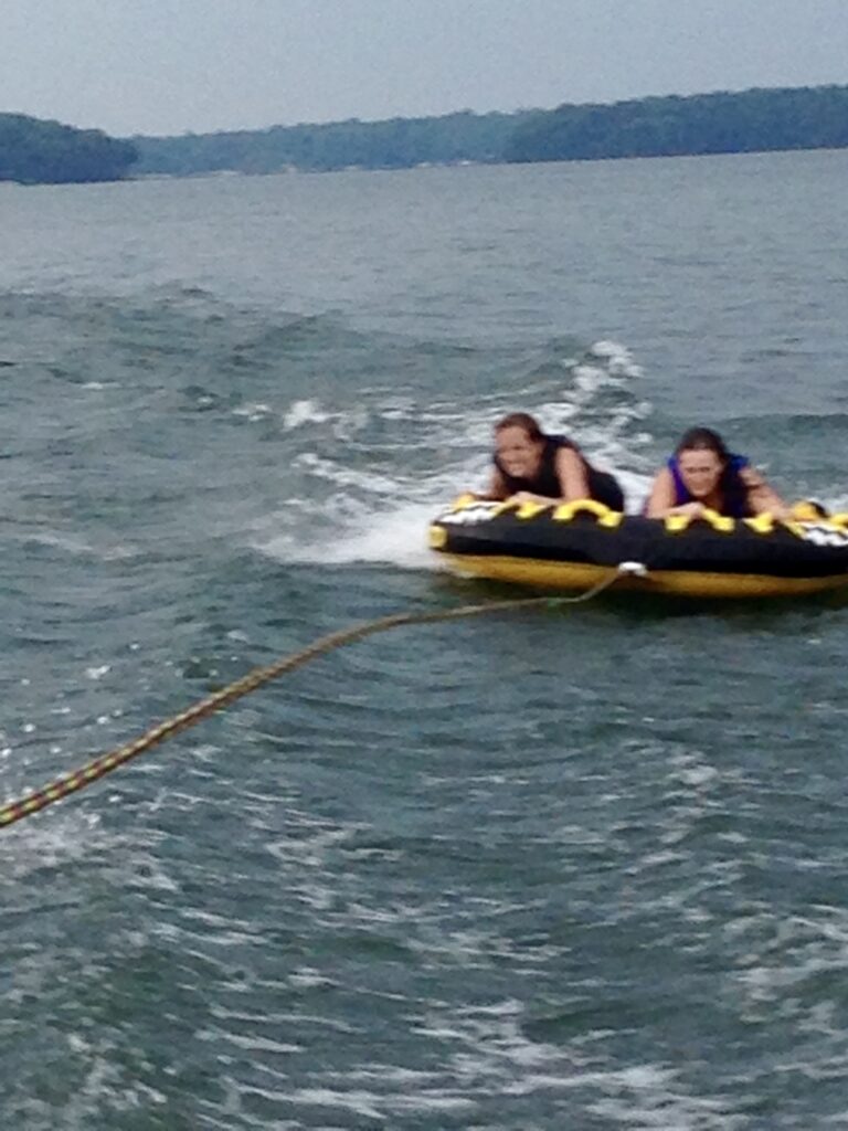 Two people wearing life jackets ride a yellow and black inflatable tube, being pulled by a boat across a large lake—a perfect choice if you’re seeking fun things to do in Charlotte. Trees line the distant shoreline under a cloudy sky.
