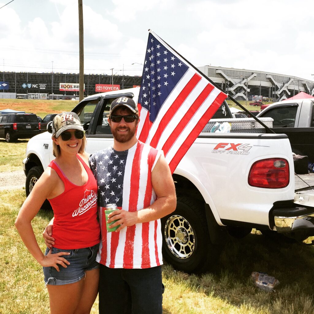 A smiling man and woman pose in front of a white pickup truck with an American flag attached—showing off their patriotic spirit at a racetrack, one of the fun things to do in Charlotte.