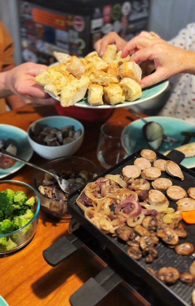 Two people share a dish of fried food over a table set with bowls of broccoli, mushrooms, and other vegetables, and a grill pan cooking sausages, onions, and carrots.