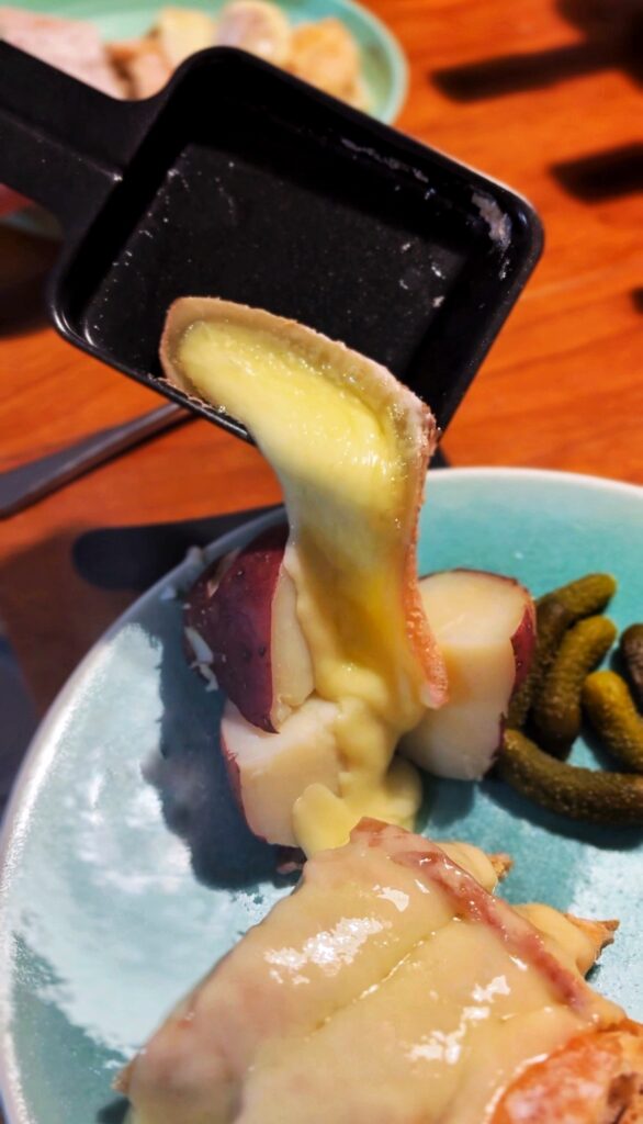 A close-up of melted cheese being poured over boiled potatoes on a blue plate, with pickles on the side.