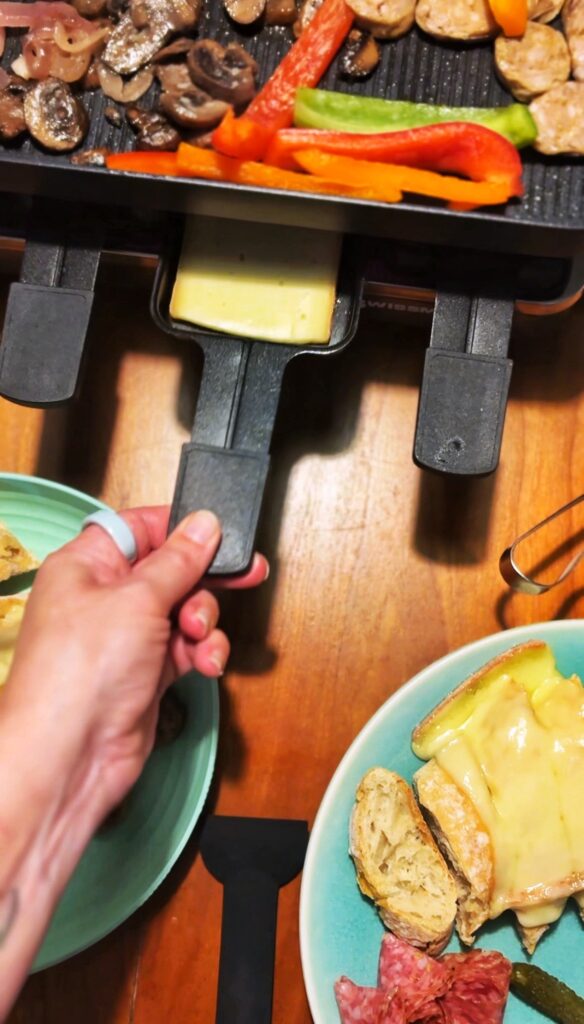 A hand pulls out a tray of melted cheese from a raclette grill, with vegetables on the grill above. Plates with bread, melted cheese, and cured meat sit on a wooden table nearby.