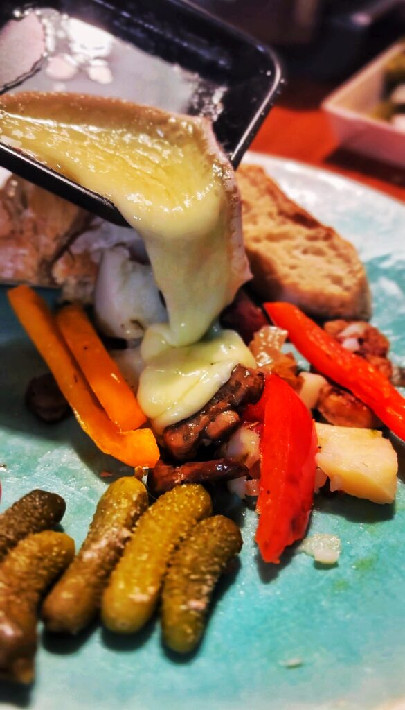 Melted cheese is being poured over roasted vegetables and bread on a turquoise plate, with several small pickles arranged in the foreground.