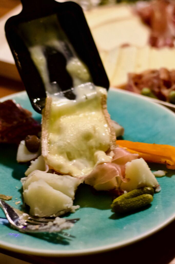 A plate with melted cheese being scraped onto prosciutto, boiled potatoes, carrots, pickles, and capers, with a fork on the side.