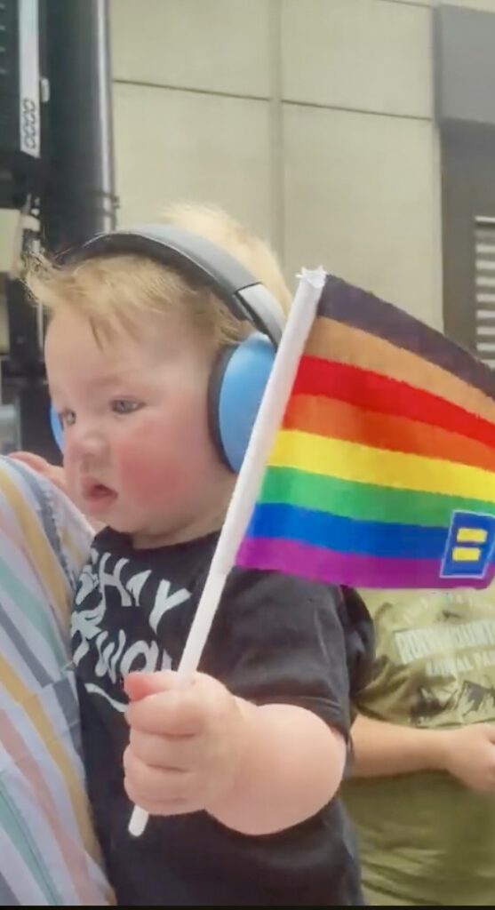 A young child wearing blue earmuffs holds a rainbow Pride flag with a yellow equal sign during an outdoor event, highlighting one of the vibrant things to do in Charlotte.