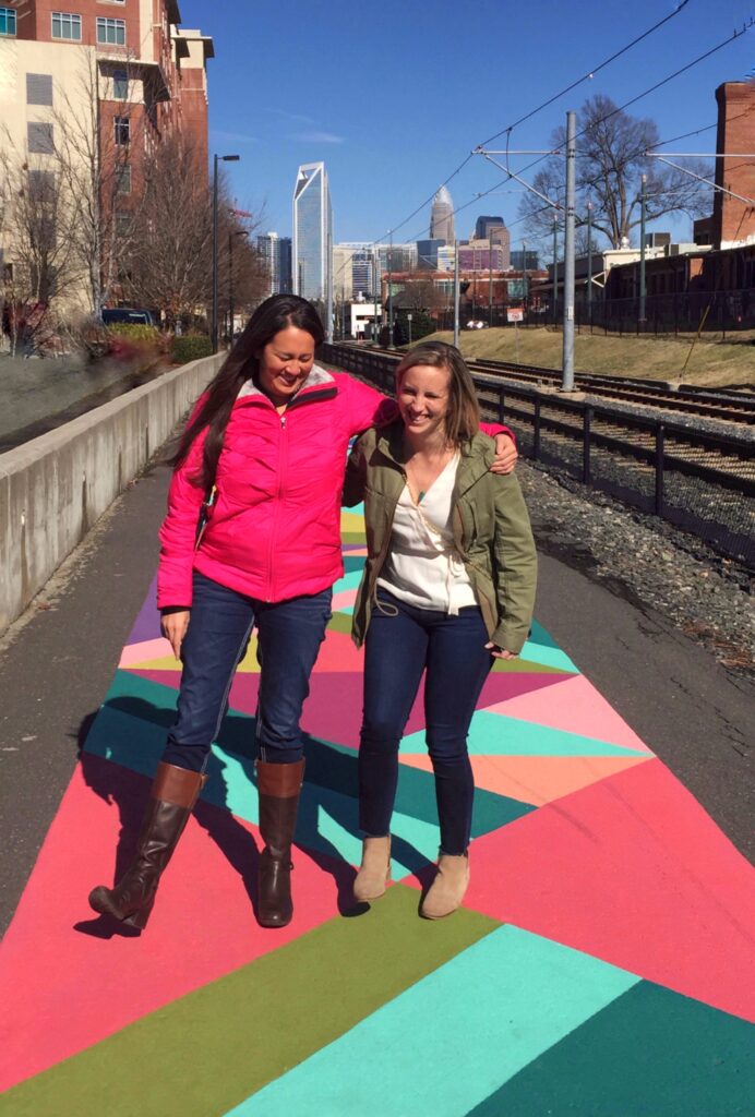 Two women stand smiling on a colorful geometric-patterned walkway near train tracks, with city skyscrapers behind them—a vibrant snapshot of the exciting things to do in Charlotte on a clear, sunny day.