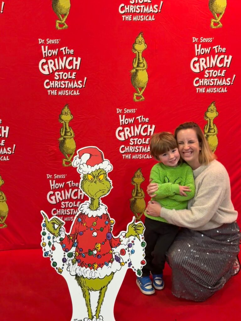 A woman and a young boy smile and pose on the red carpet in front of a How the Grinch Stole Christmas! The Musical backdrop, one of the festive things to do in Charlotte, next to a cardboard cutout of the Grinch in a Santa hat.