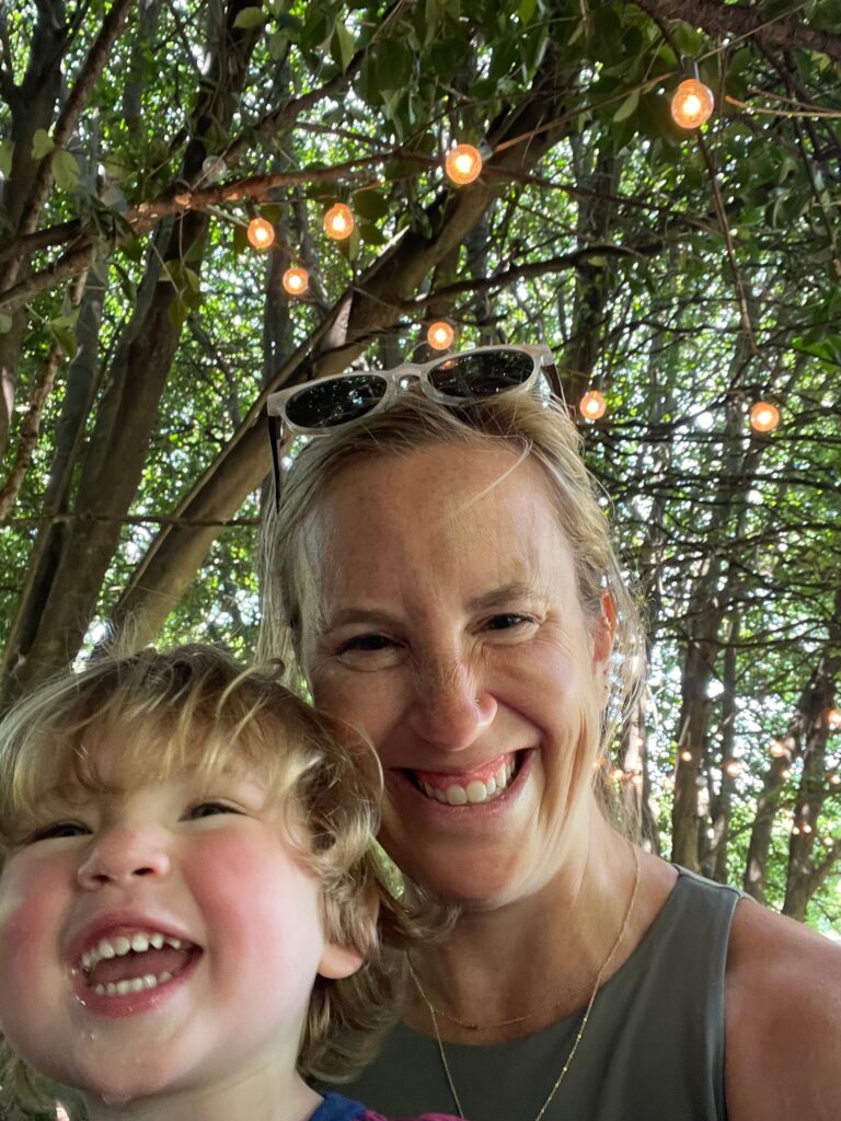 A woman and a young child smile happily under a canopy of trees with string lights glowing above them—a perfect scene for those seeking fun things to do in Charlotte in a sunlit outdoor setting.