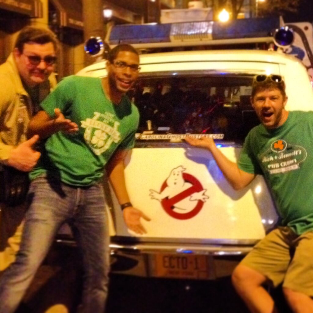 Three men pose excitedly in front of a white car featuring the Ghostbusters logo and ECTO-1 plate at night—one fun snapshot from things to do in Charlotte. Two wear green T-shirts, while one sports glasses and a tan jacket.