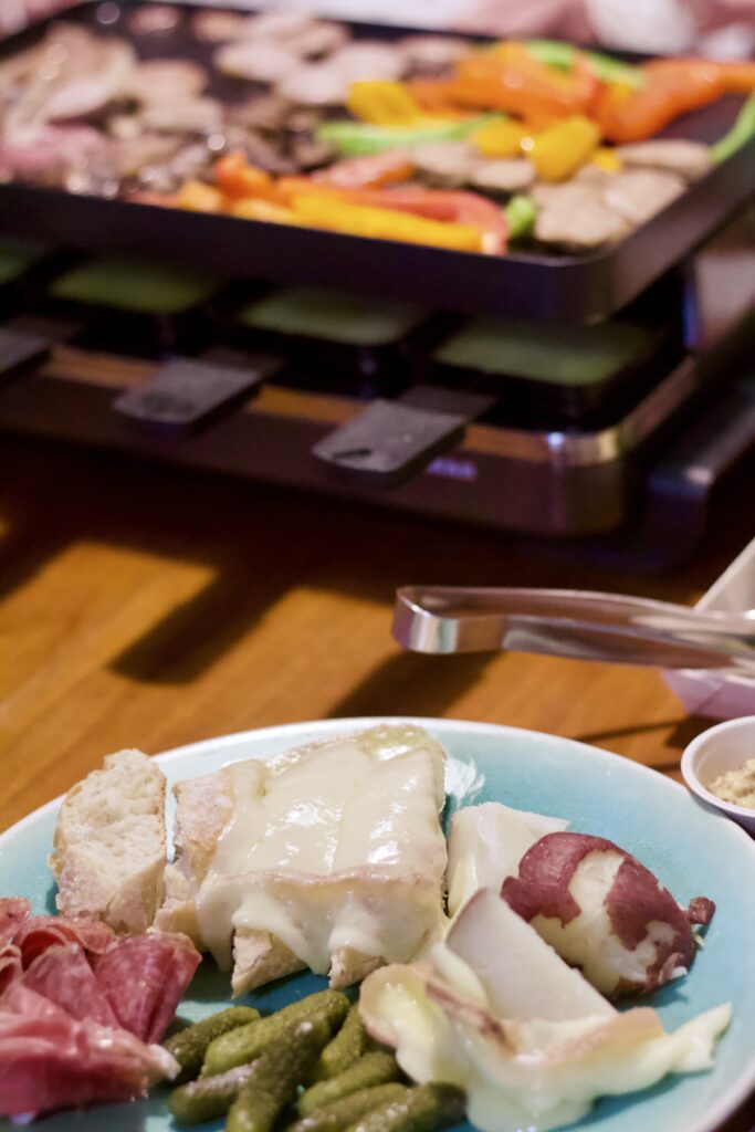 A plate with sliced bread, melted cheese, pickles, and cured meats sits in front of a raclette grill loaded with melting cheese, vegetables, and meats—perfect for a cozy raclette dinner party.