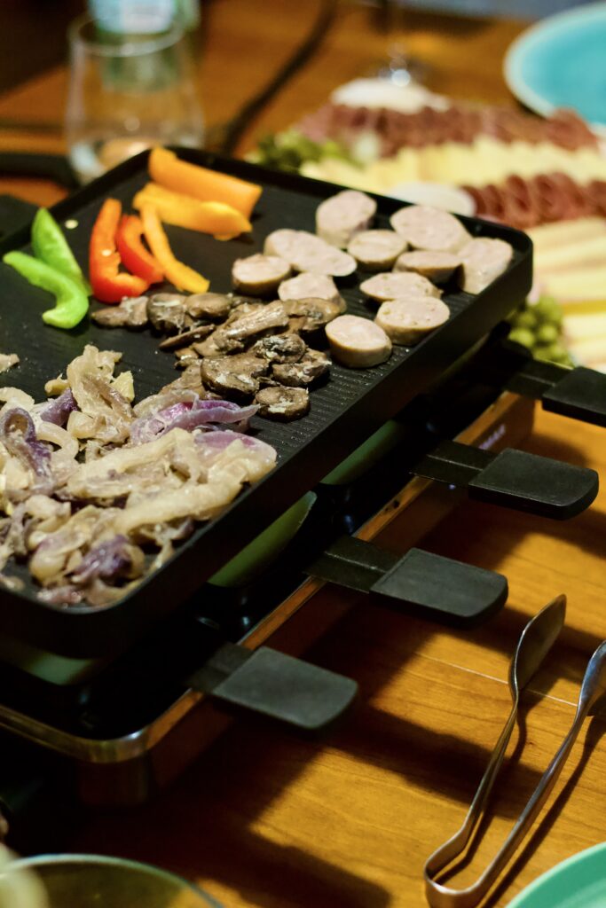A tabletop grill sizzles with sliced sausages, bell peppers, mushrooms, and onions at a lively raclette dinner party, serving utensils and plates set around the wooden table.