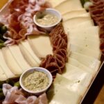 A charcuterie board with neatly arranged slices of cheese, folded cured meats, pickles, and two small cups of grainy mustard on a wooden surface—perfect for kicking off a cozy raclette dinner party.
