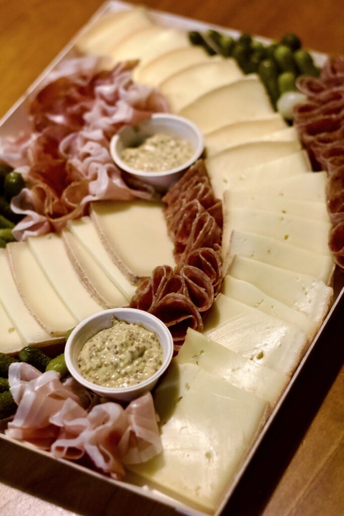 A charcuterie board with neatly arranged slices of cheese, folded cured meats, pickles, and two small cups of grainy mustard on a wooden surface—perfect for kicking off a cozy raclette dinner party.