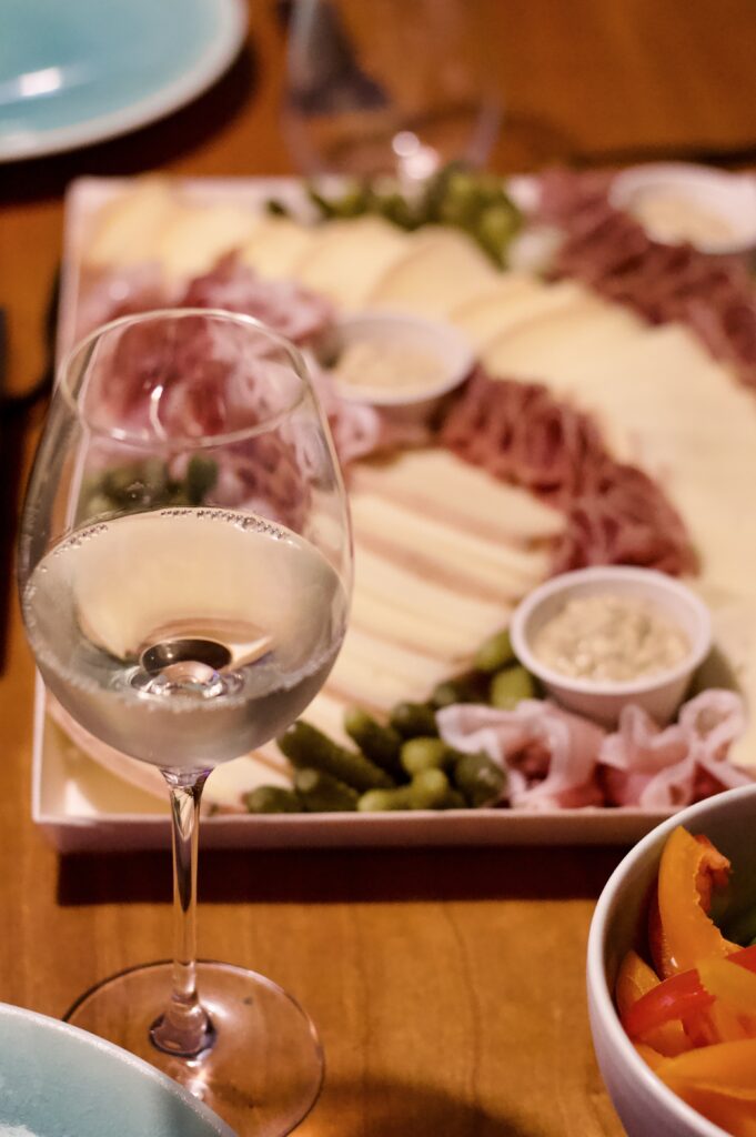 A glass of white wine on a wooden table, with a platter of assorted cheeses, meats, pickles, and dips—perfect for a cozy raclette dinner party—plus a bowl of sliced red and yellow peppers in the foreground.