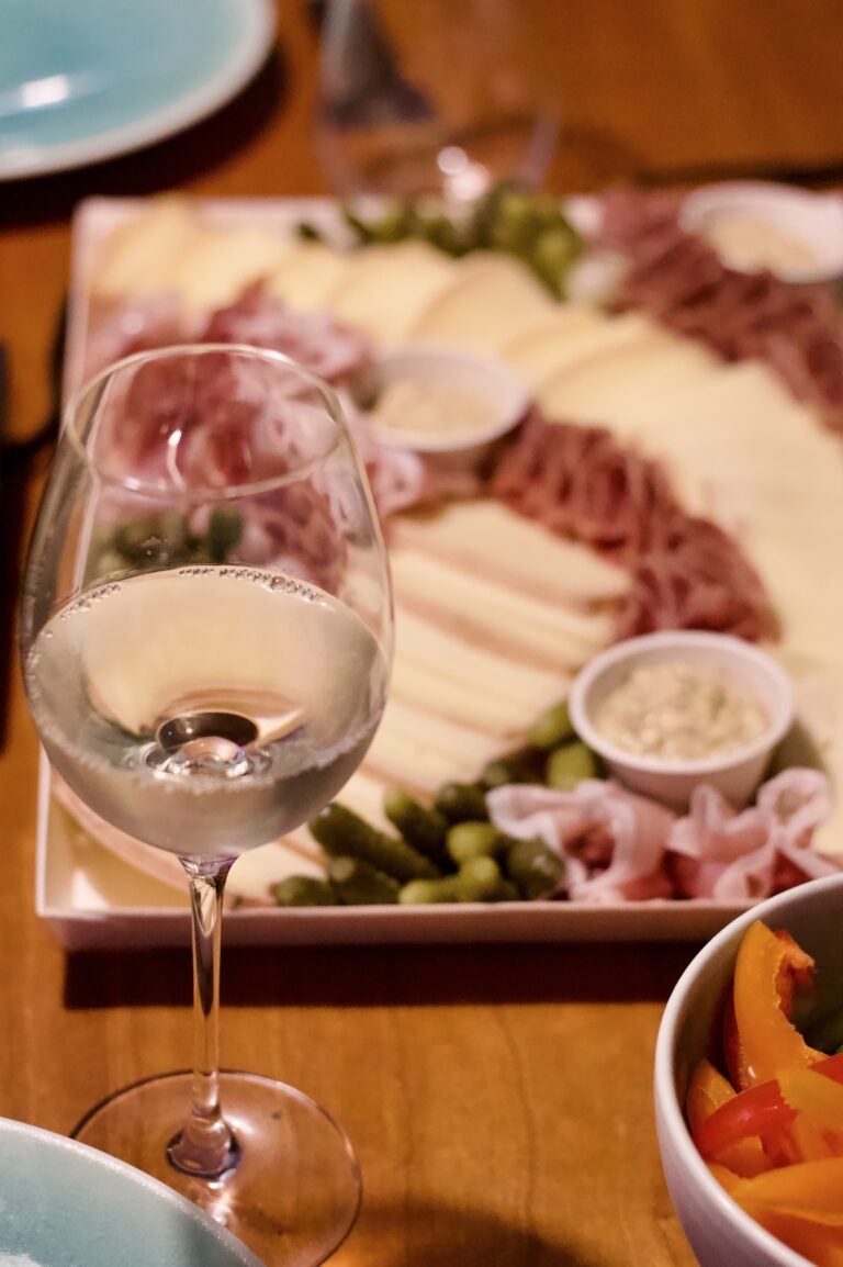 A glass of white wine on a wooden table, with a platter of assorted cheeses, meats, pickles, and dips—perfect for a cozy raclette dinner party—plus a bowl of sliced red and yellow peppers in the foreground.