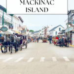 A busy street on Mackinac Island with horse-drawn carriages, historic buildings, and people walking. Text overlay reads: “Mackinac Island Day Trip: One Day Itinerary for Visiting Mackinac Island.”.