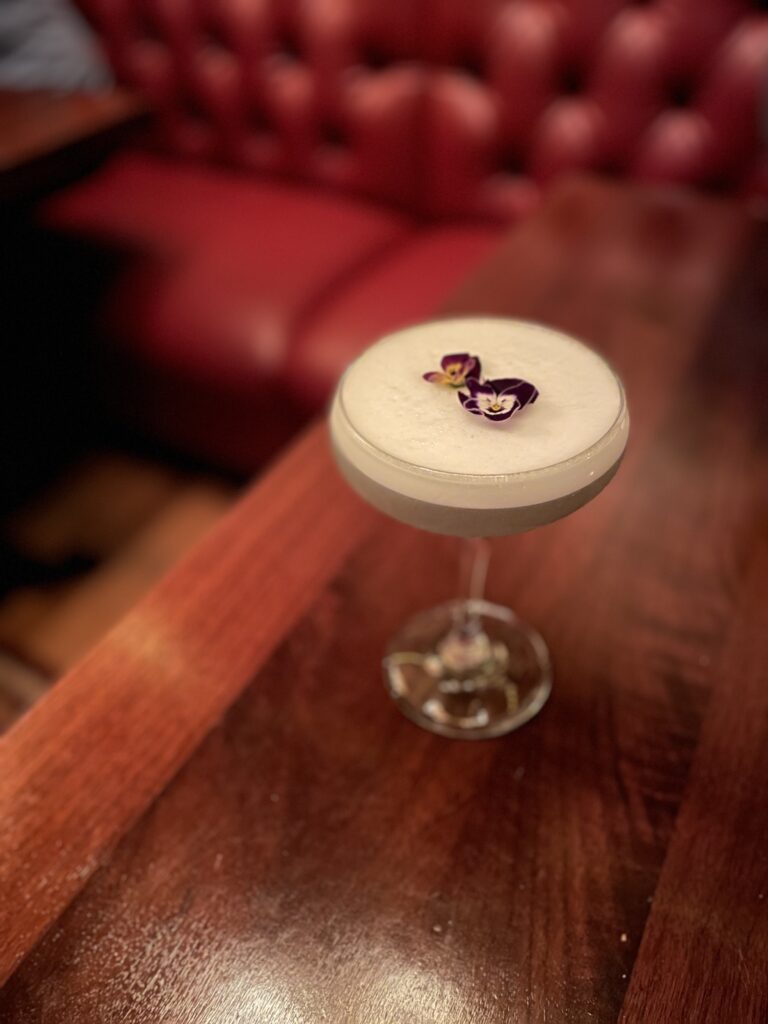 A frothy cocktail in a coupe glass is garnished with edible purple flowers, sitting on a polished wooden table with a red tufted leather booth in the background.