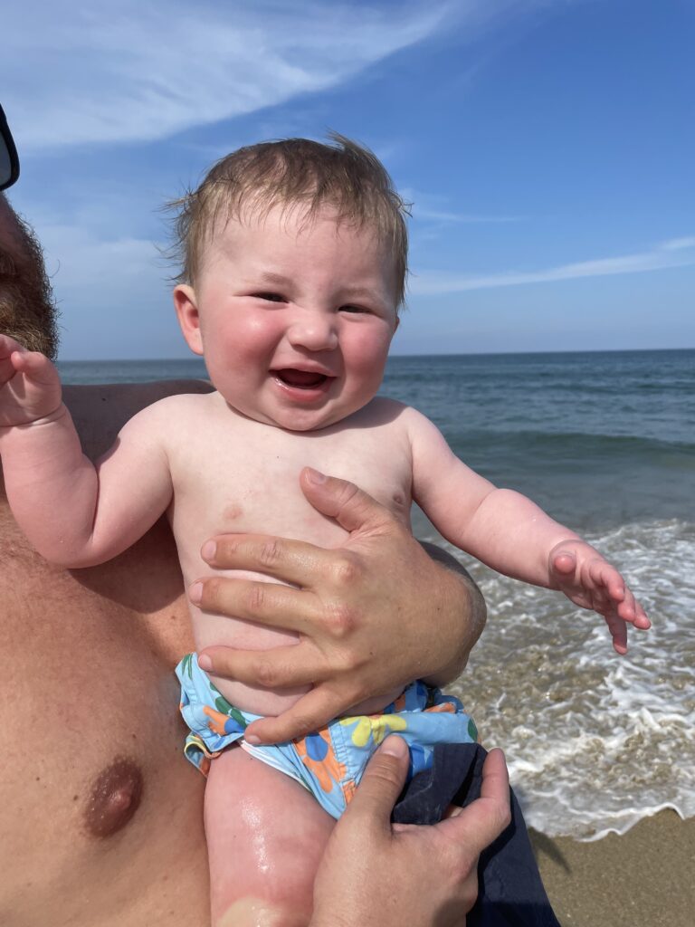 A smiling baby in patterned swim trunks is held by an adult at the beach, with the ocean and blue sky in the background. The baby looks happy, with wet hair and a hand raised in the air.