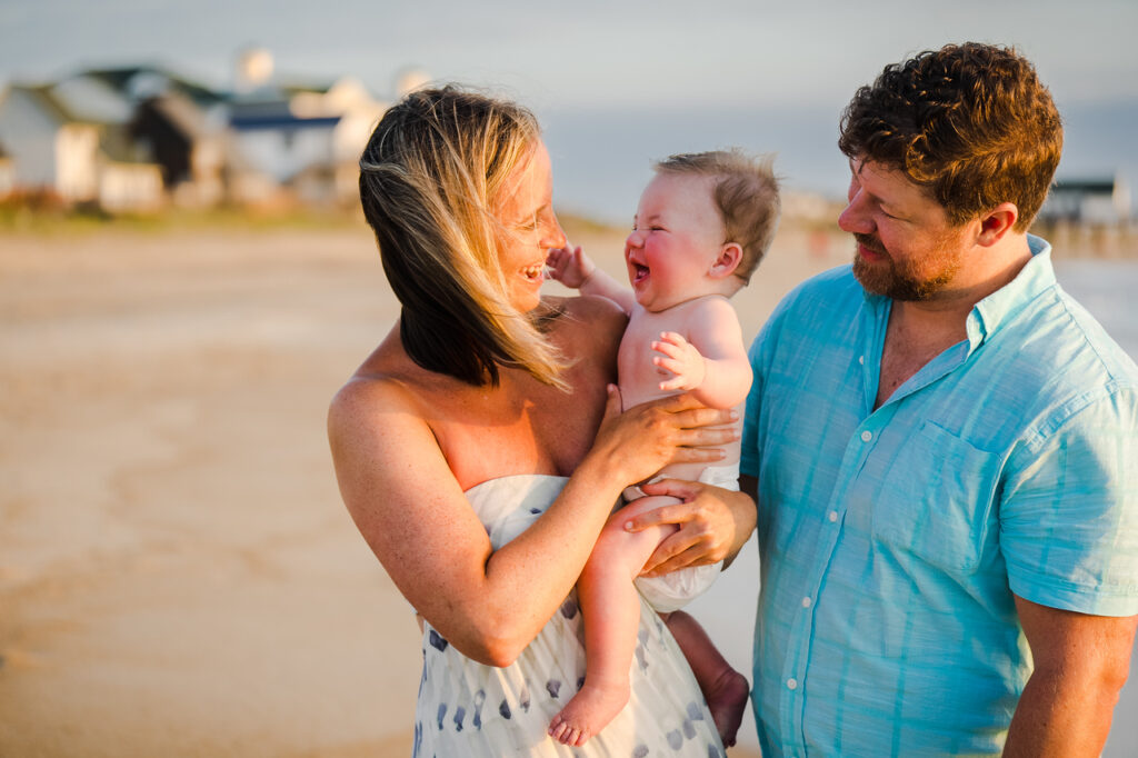 A woman and man smile and hold a laughing baby at the beach. The woman wears a strapless dress, the man wears a light blue shirt, and the background shows sand, water, and blurry beach houses.