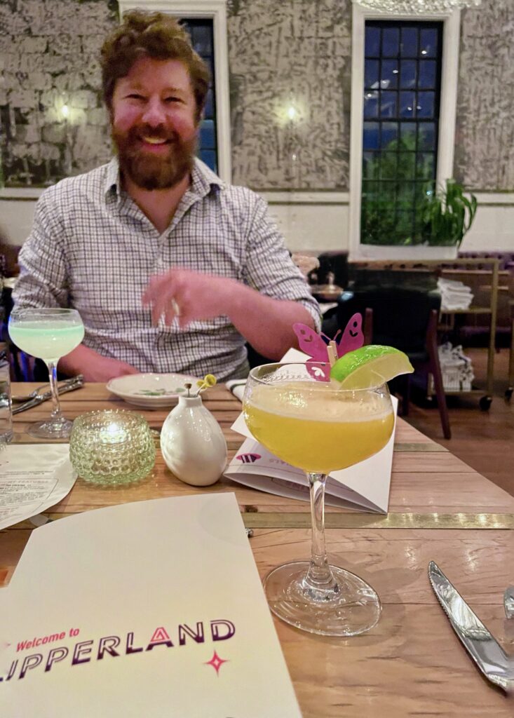 A smiling man with a beard sits at a restaurant table with two colorful cocktails; one has a purple butterfly decoration and a lime. A menu, candle, and cutlery are on the wooden table in front of him.