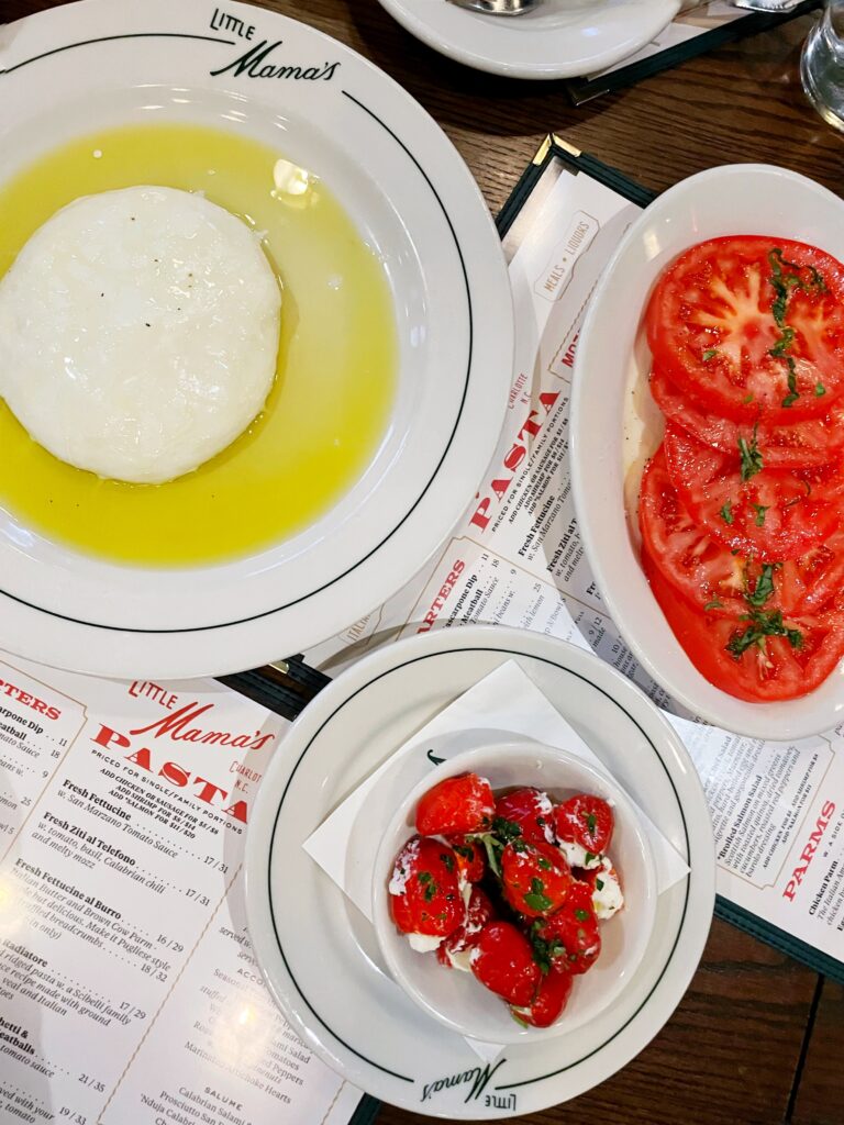 Plates of fresh mozzarella in olive oil, sliced tomatoes with herbs, and cherry tomatoes with cheese are arranged on a table set with menus at Little Mama’s restaurant.
