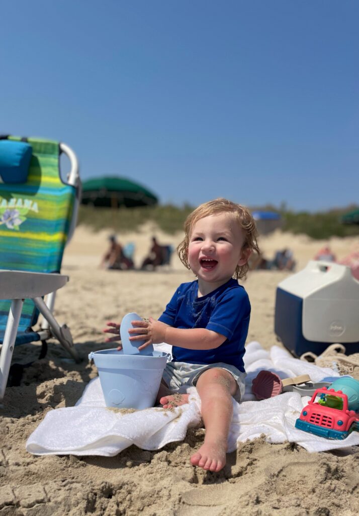 A smiling toddler in a blue shirt sits on a beach towel playing with a bucket and shovel. A colorful toy truck, beach chair, and cooler are nearby. People and umbrellas are in the background under a clear blue sky.