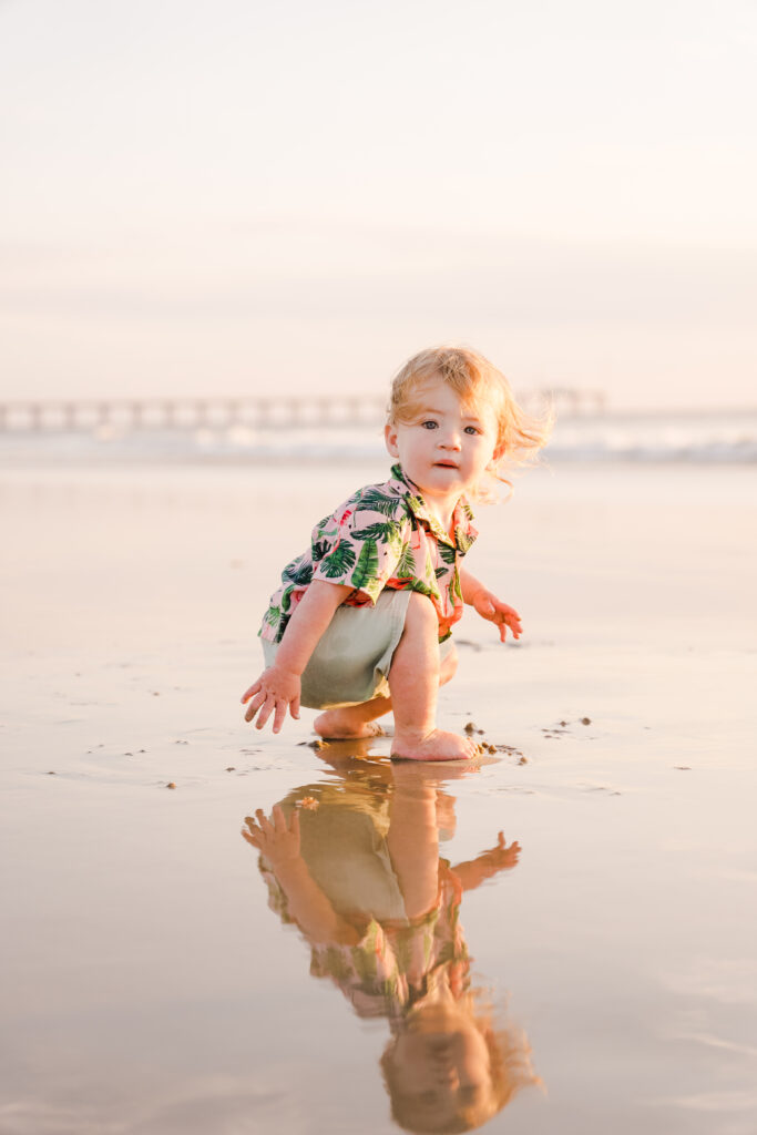 A young child with blonde hair squats on a sandy beach, reflected in the wet sand. They wear a colorful shirt and look toward the camera. The ocean and a distant pier are visible in the background.