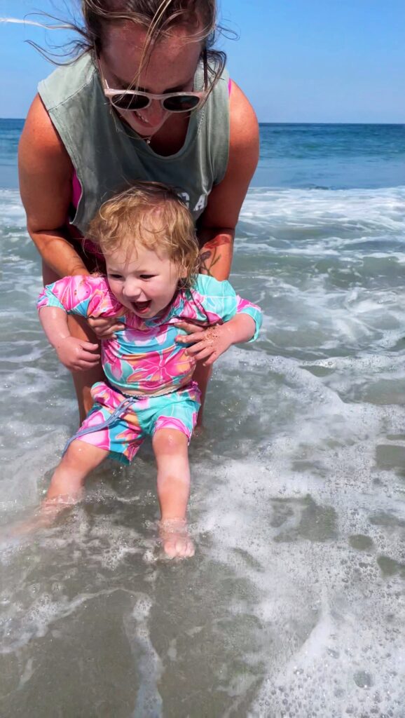 An adult holds a smiling toddler in a colorful swimsuit above shallow ocean waves, helping her play in the water on a sunny day.