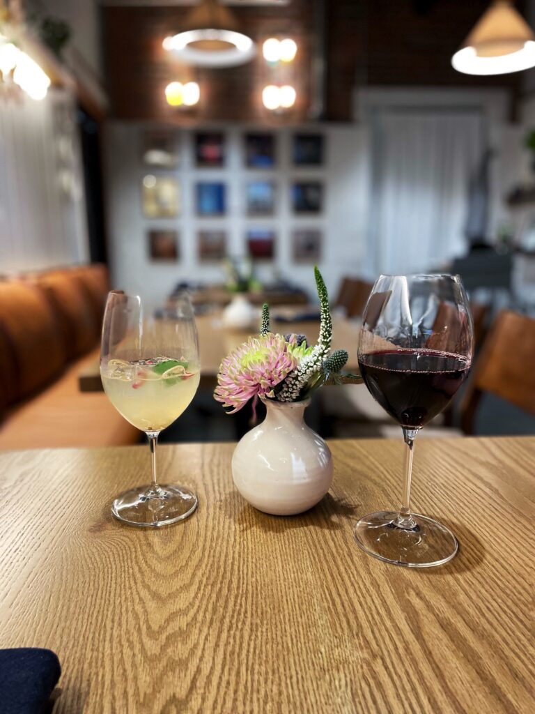Two wine glasses, one with white wine and the other with red wine, sit on a wooden table next to a small white vase with pink flowers in a cozy, softly-lit restaurant.