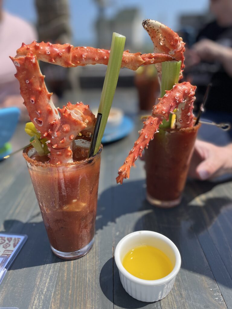 A Bloody Mary cocktail garnished with large king crab legs and celery sticks on an outdoor table, with a small ramekin of melted butter nearby. Another similar drink and a blurred figure are in the background.