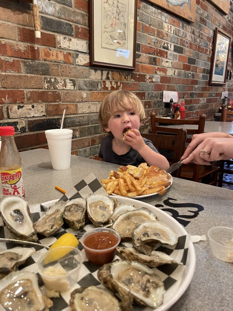 A young child sits at a restaurant table eating French fries and a sandwich, while a plate of raw oysters with sauces and lemon is in the foreground. The background features a brick wall with framed art.