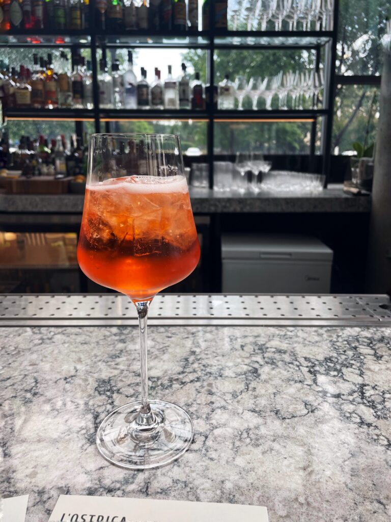 A glass of orange-colored cocktail with ice sits on a marble bar counter. Shelves with bottles and glassware are visible in the background, and natural light streams in through large windows.