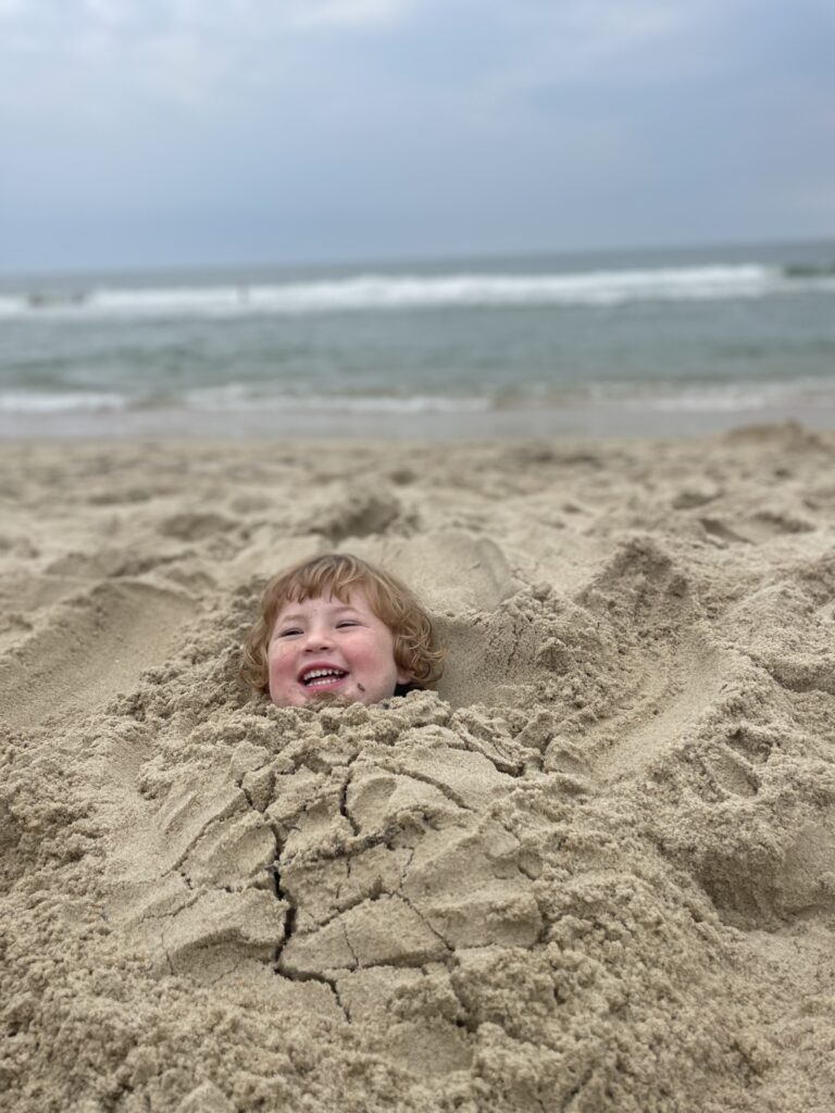 A young child, buried up to their neck in sand, smiles joyfully at the beach with waves and a cloudy sky in the background.