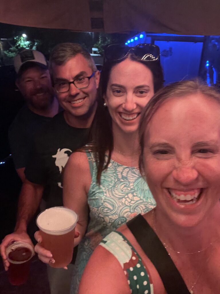 Four smiling adults stand closely together indoors, holding drinks. The woman in front is taking the selfie, and all appear to be enjoying a casual, fun night out. The background is dimly lit with some blue lights visible.