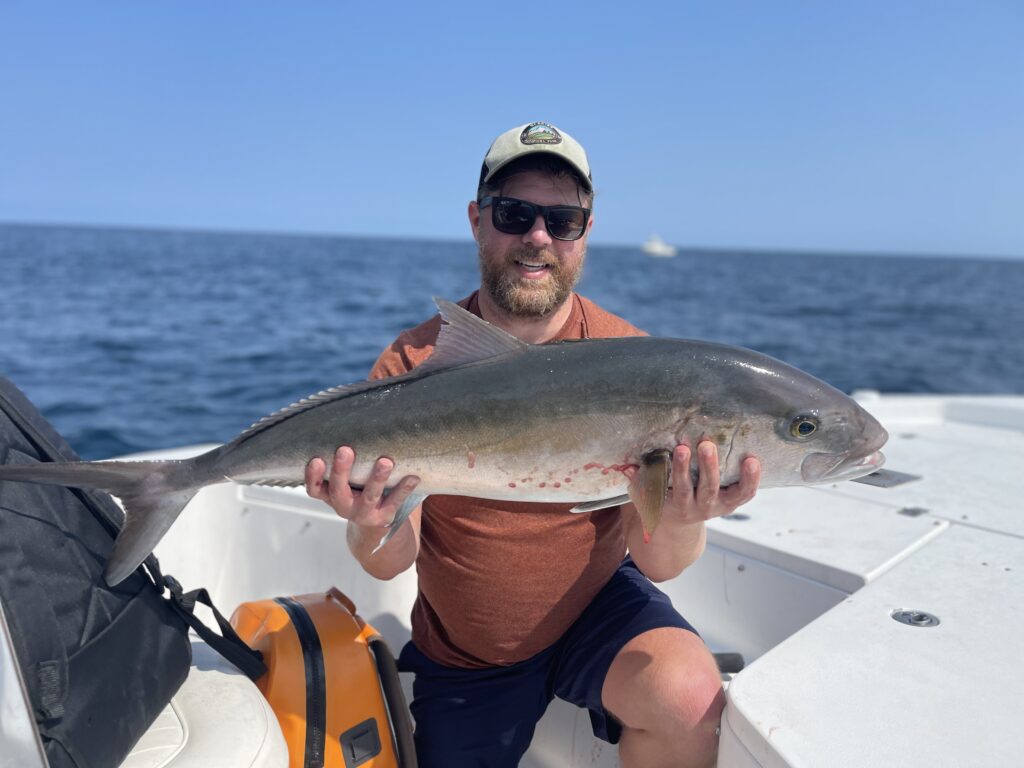 A man wearing sunglasses, a cap, and a reddish shirt sits on a boat holding a large fish with both hands. The ocean and a distant boat are visible in the background under a clear blue sky.