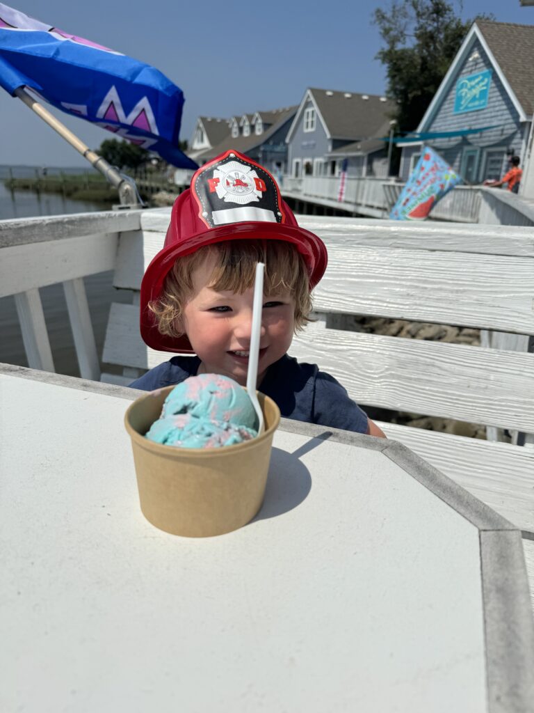 A young child wearing a red toy firefighter hat smiles behind a paper cup of colorful ice cream at an outdoor table, with seaside cottages and bright flags in the background.