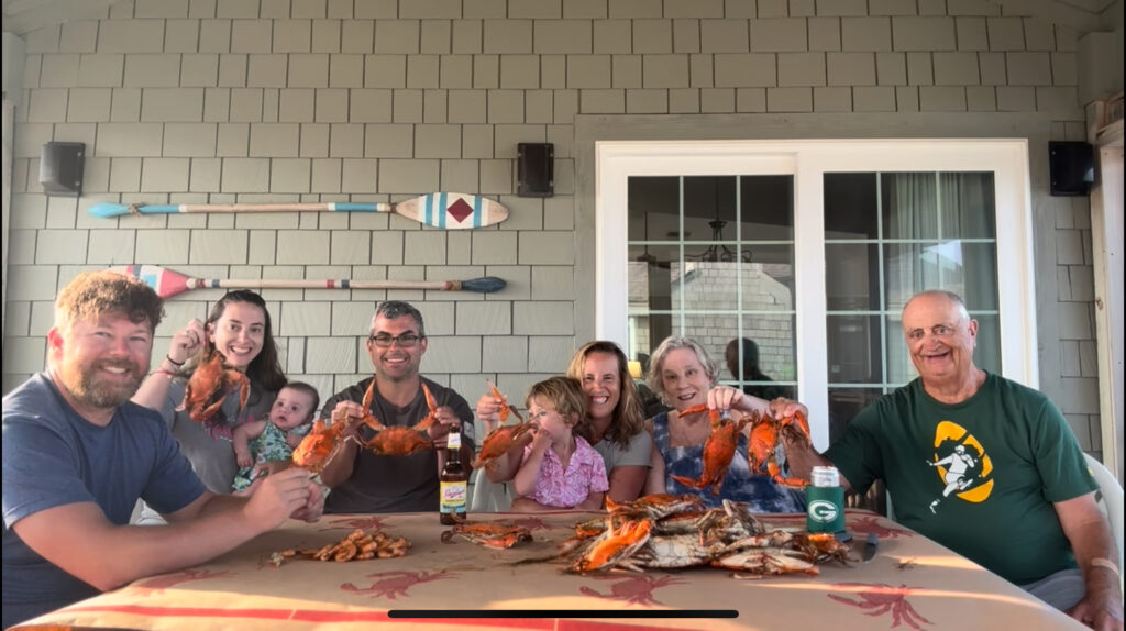A group of six adults and two children sit around a table covered with steamed crabs and drinks, smiling and holding up crabs, enjoying a meal on a patio decorated with colorful paddles.
