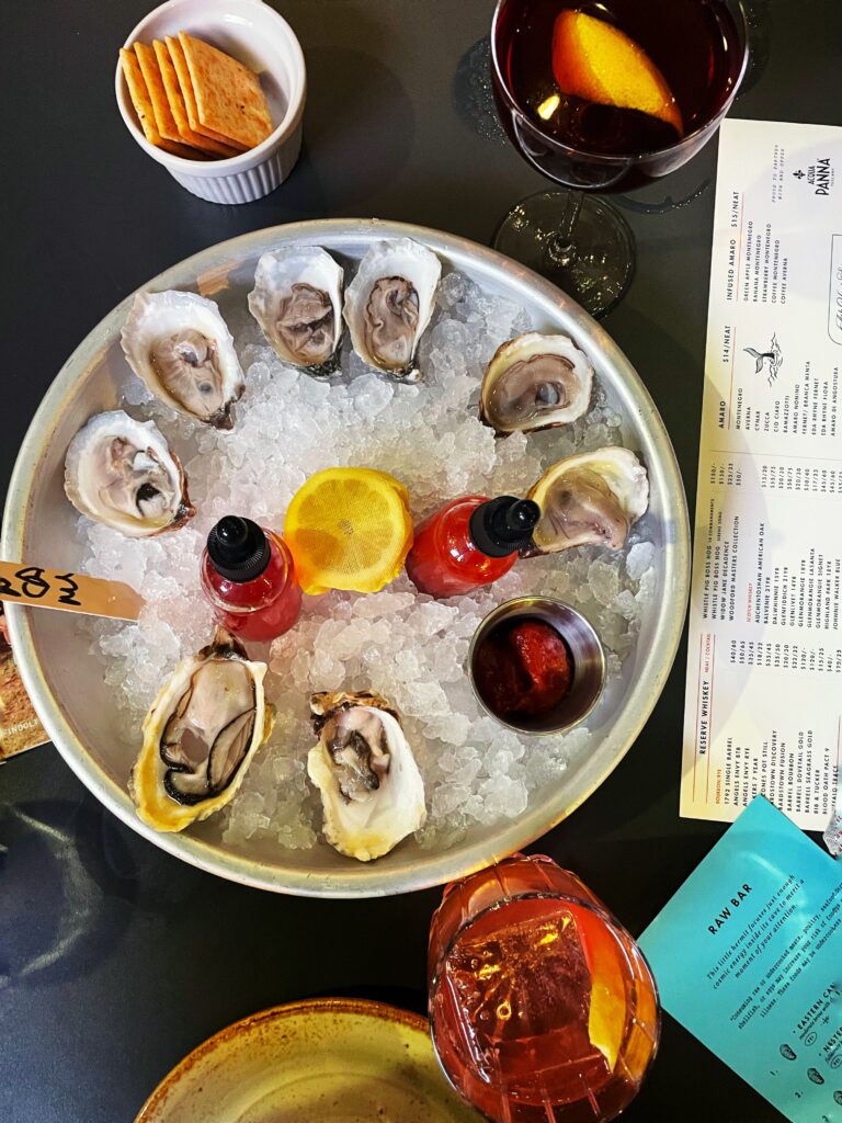 A plate of oysters on ice with lemon slices and red sauce bottles, surrounded by drinks, a small bowl of crackers, and restaurant menus on a dark tabletop.