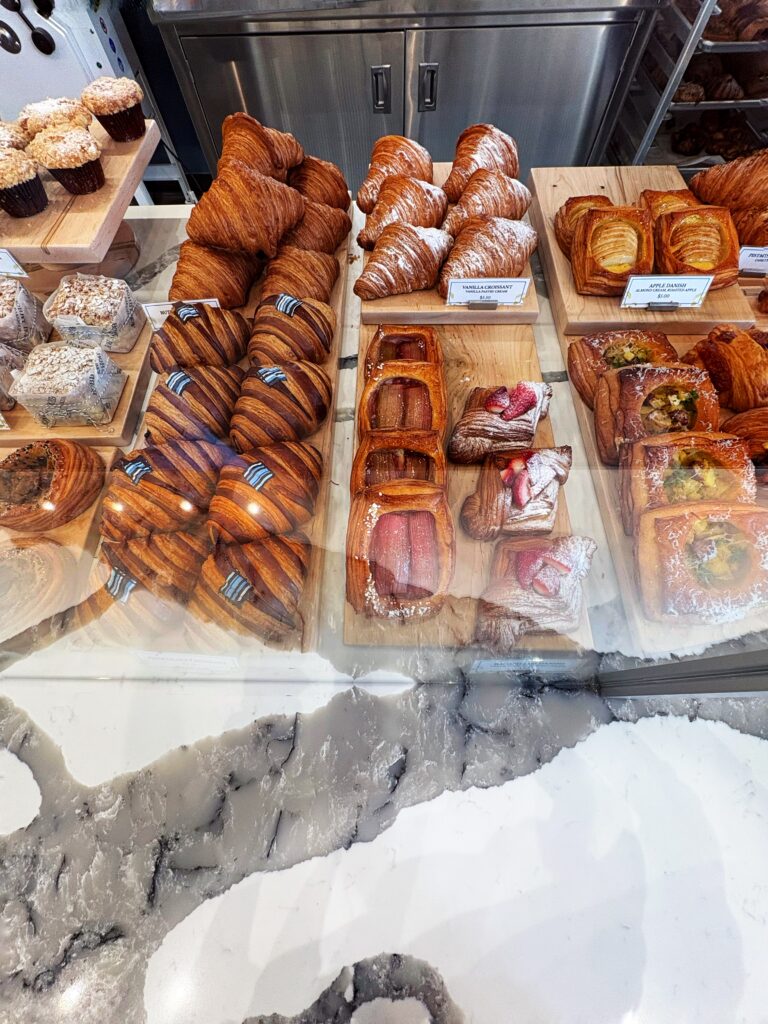 A bakery display case filled with assorted pastries, including croissants, chocolate croissants, and fruit-topped danishes, all arranged neatly on wooden boards.