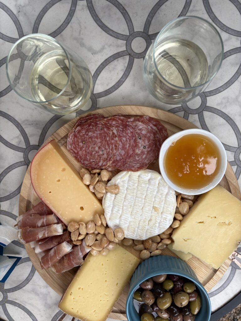 A round wooden board with assorted cheeses, salami, prosciutto, marcona almonds, green olives in a bowl, and a small dish of apricot jam. Two glasses of white wine are beside the board on a patterned surface.