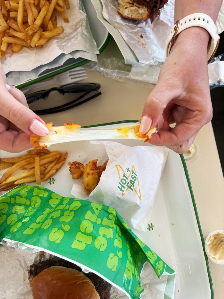 A person pulls apart a mozzarella stick with melted cheese stretching between their hands, surrounded by fries, a burger, and food wrappers on a table.