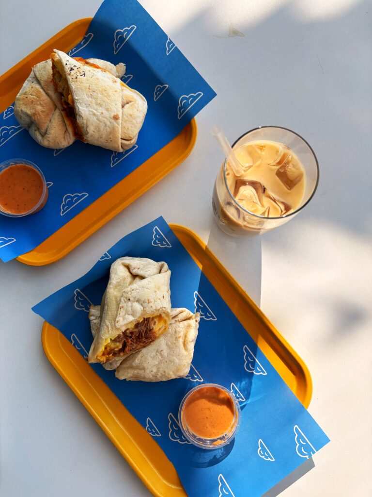 Two breakfast burritos on yellow trays lined with blue cloud-patterned paper, each with a cup of dipping sauce. A glass of iced coffee with a straw sits to the side on a white surface in bright natural light.