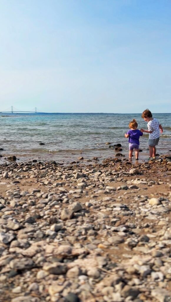 Two young children stand on a rocky beach by the shore, looking out at the water. A bridge is visible in the distance under a clear blue sky.
