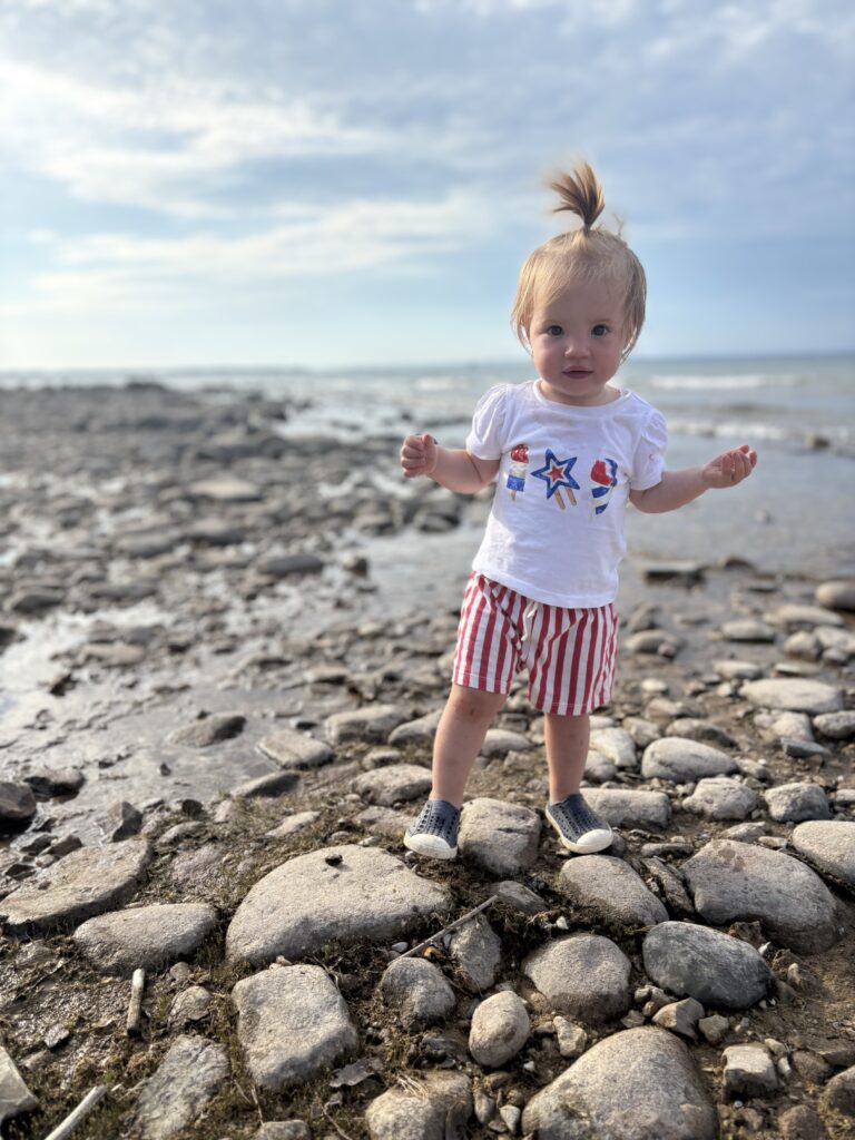 A toddler with blond hair in a high ponytail stands on rocky ground near a body of water, wearing a white shirt with stars and red-striped shorts under a partly cloudy sky.