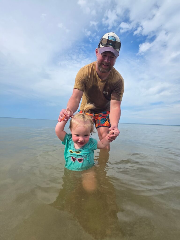 A man in a cap and sunglasses holds a smiling toddler’s hands as they stand together in the shallow water at the beach under a partly cloudy sky. The child wears a turquoise shirt and looks happy.