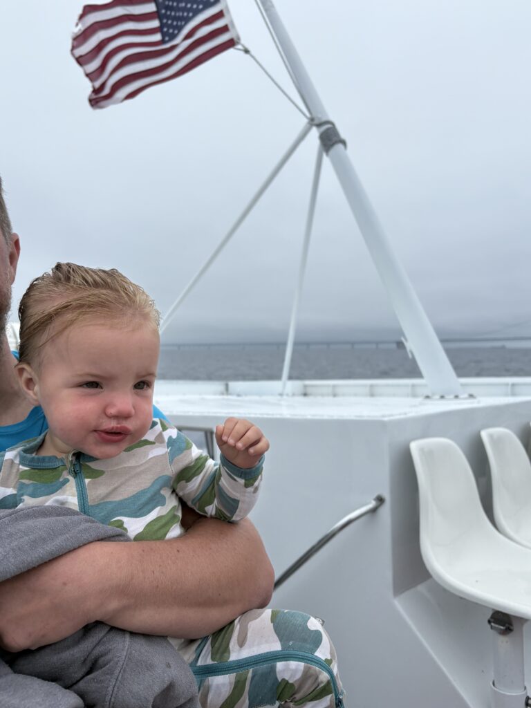 A young child in a camouflage-patterned outfit is held by an adult on a boat. The child’s hair is windswept, and an American flag waves in the background against a cloudy, overcast sky. Empty white chairs are visible nearby.