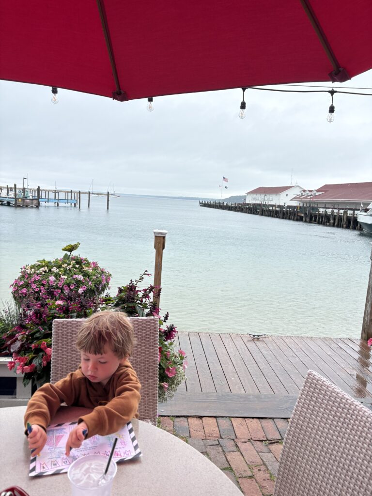 A young child sits at an outdoor table under a red umbrella, coloring on paper. Behind them is a wooden deck, pink flowers, calm water, docks, and boats on a cloudy day.