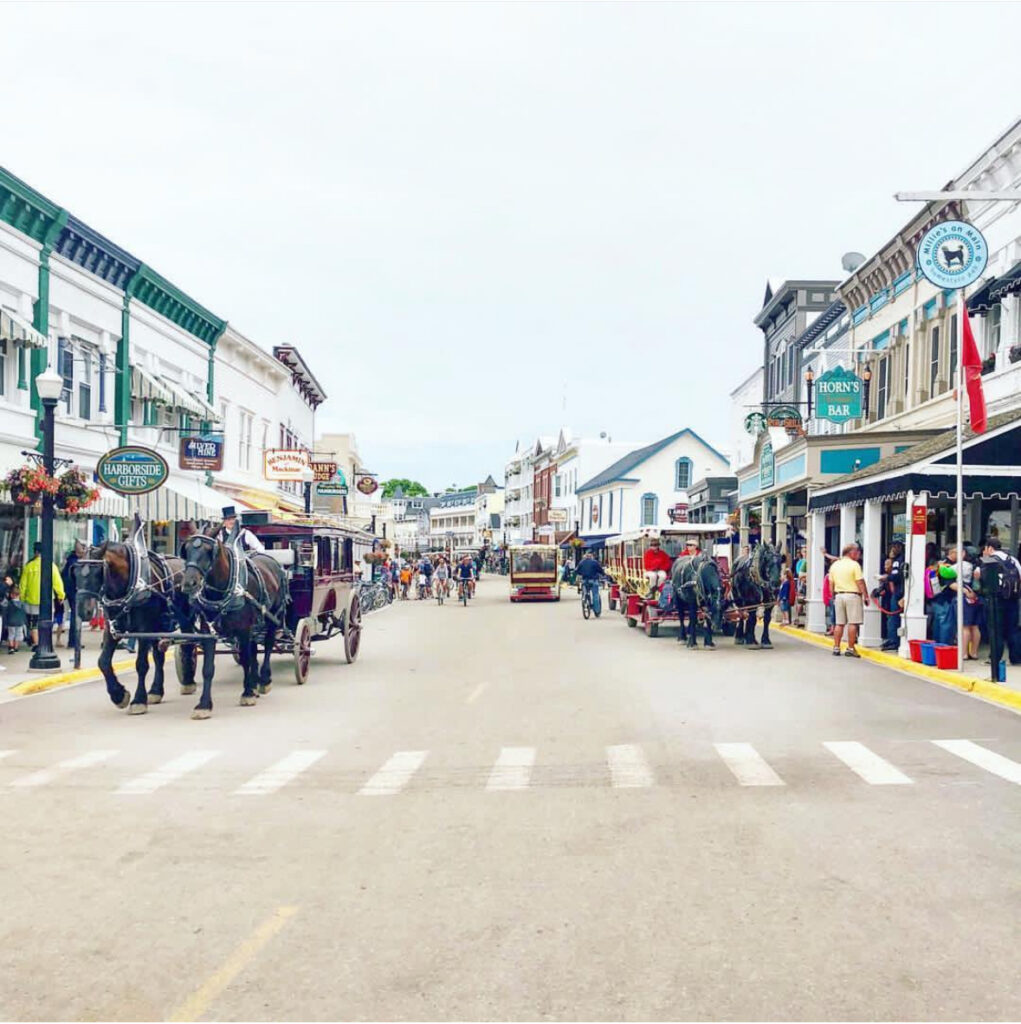 A busy street scene on Mackinac Island, Michigan, with horse-drawn carriages, people walking, and historic buildings lining both sides of the street under a bright sky.