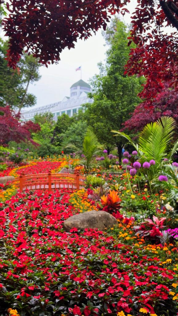 A lush garden filled with vibrant red, orange, and purple flowers surrounds a small orange footbridge, with a white building and trees visible in the background.