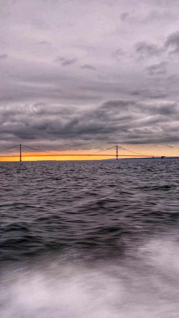 A long suspension bridge stretches across choppy water beneath a dramatic, cloudy sky at sunset. The horizon glows with a strip of warm orange light, contrasting with the dark clouds and water.